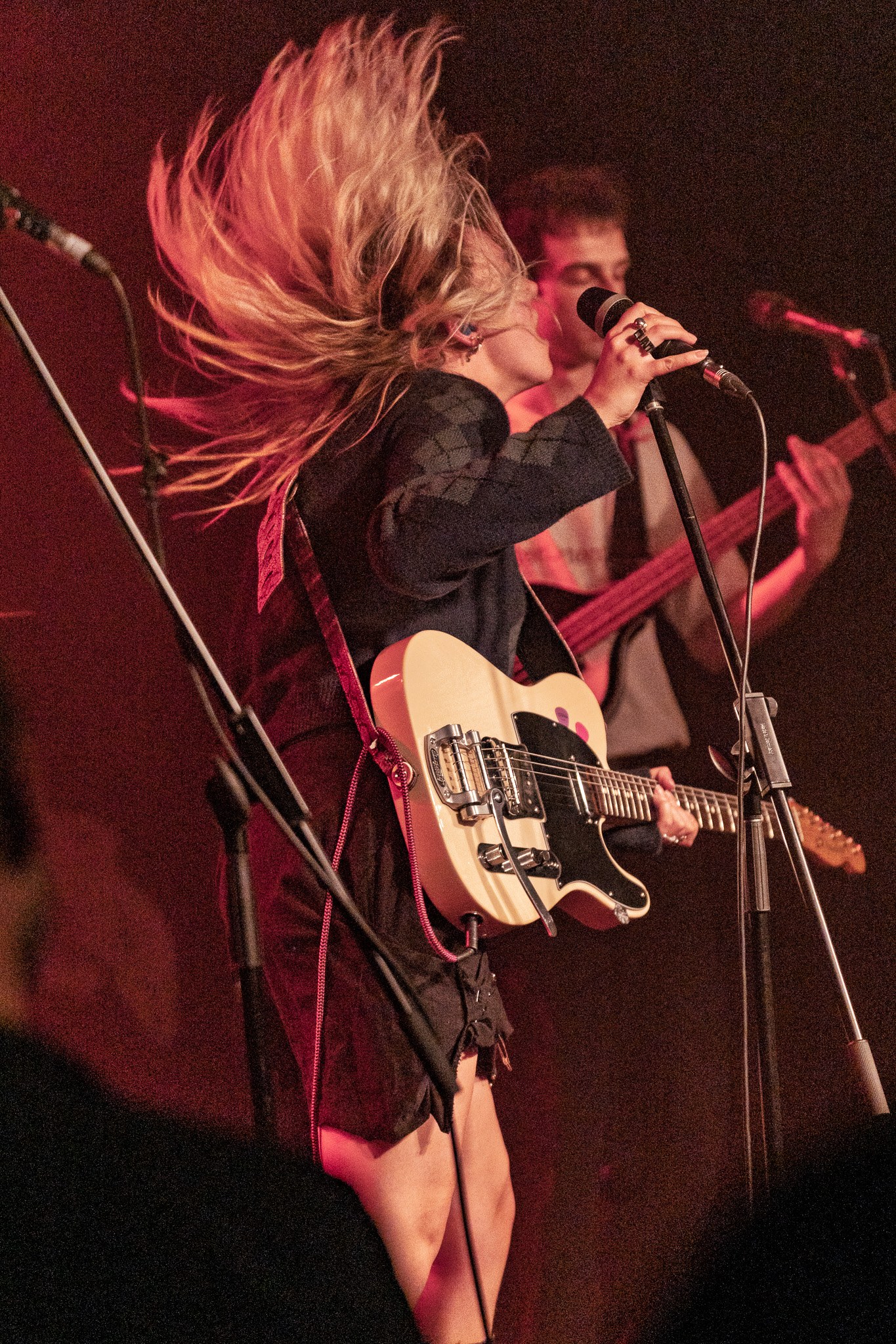 A singer with hair in motion performing with an electric guitar under red stage light.
