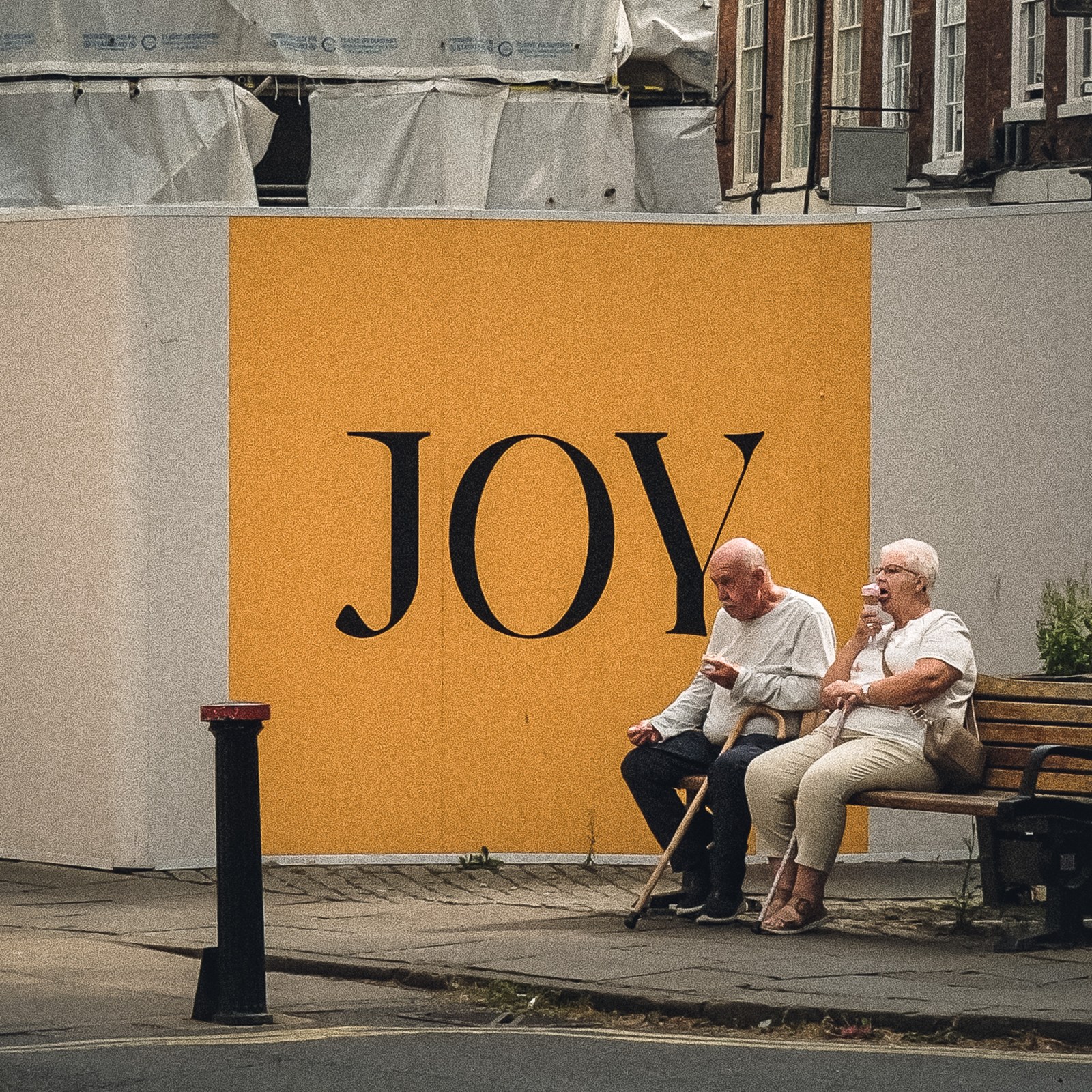 Two people on a bench beside a yellow wall with the word JOY.